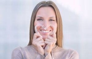 Woman with perfect smile holding invisible dental aligners bent in a shape of heart in front of her face.