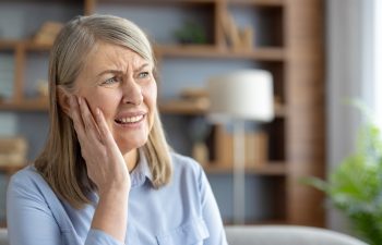 Elderly Caucasian woman suffering from otitis, touching her ear in pain, sitting in a modern living room. She looks distressed and worried about her earache.